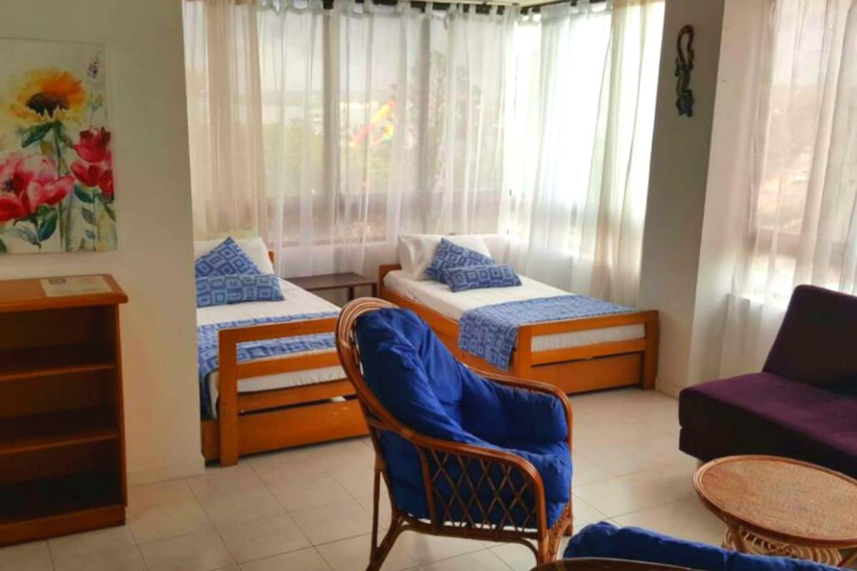 A room with two single beds, a wicker chair, a purple sofa, and a small table.