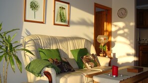 Una sala de estar con un sofá color beige, cojines verdes, una mesa de centro de madera con una vela roja, e impresiones botánicas enmarcadas en la pared.