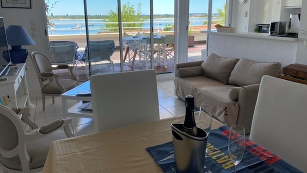 Ein modernes Wohnzimmer mit Sofa, Esstisch und Stühlen. Eine große Schiebeglastür führt zu einer Außenpatio mit Blick auf ein Gewässer.