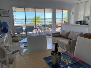 En moderne stue med sofa, spisebord og stole. En stor skydedør fører ud til en udendørs terrasse med udsigt over vandet.