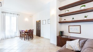 Una sala de estar con un sofá marrón, una mesa de comedor con sillas, una zona de cocina y un pasillo que lleva a una puerta.