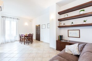 A living room with a brown sofa, a dining table with chairs, a kitchen area, and a hallway leading to a door.