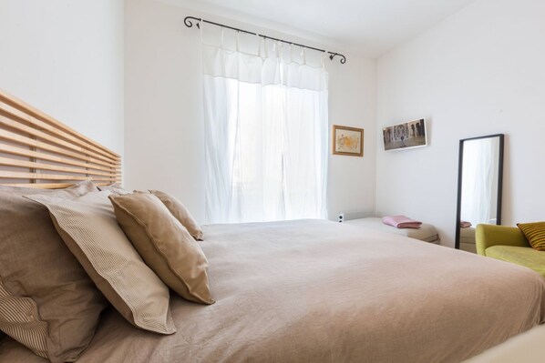Un dormitorio con una cama grande, cabecera de madera, ventana con cortinas traslúcidas, mesita de noche con lámpara y una silla.