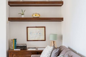 A living room with a wooden shelf, a framed abstract painting, a lamp, and a sofa with cushions.