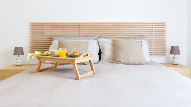 Una cama bien hecha con cabecera de madera, una mesita de noche con lámpara y una bandeja con alimentos para el desayuno.