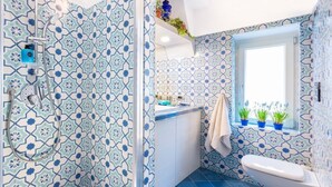 Un baño con azulejos estampados, ducha, inodoro y ventana con plantas.