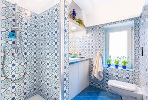 A bathroom with patterned tiles, a shower, a toilet, and a window with plants.