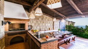 A rustic outdoor kitchen with a brick fireplace, hanging pendant lights, and a large dining table. - MyExclusiveItaly - Luxury Villa with breathtaking Seaview,pool,BBQ (Sorrento)