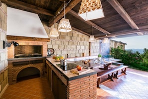 A rustic outdoor kitchen with a brick fireplace, hanging pendant lights, and a large dining table. - MyExclusiveItaly - Luxury Villa with breathtaking Seaview,pool,BBQ (Sorrento)