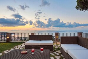A wicker sofa with cushions, a table with two glasses of red drink, and a view of the sea and cityscape.