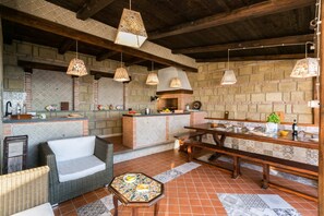 A rustic outdoor kitchen with a brick fireplace, hanging pendant lights, and a wooden bench.