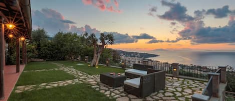 Une terrasse avec un sol en pierre, du mobilier d’extérieur et une vue sur la mer au coucher du soleil.