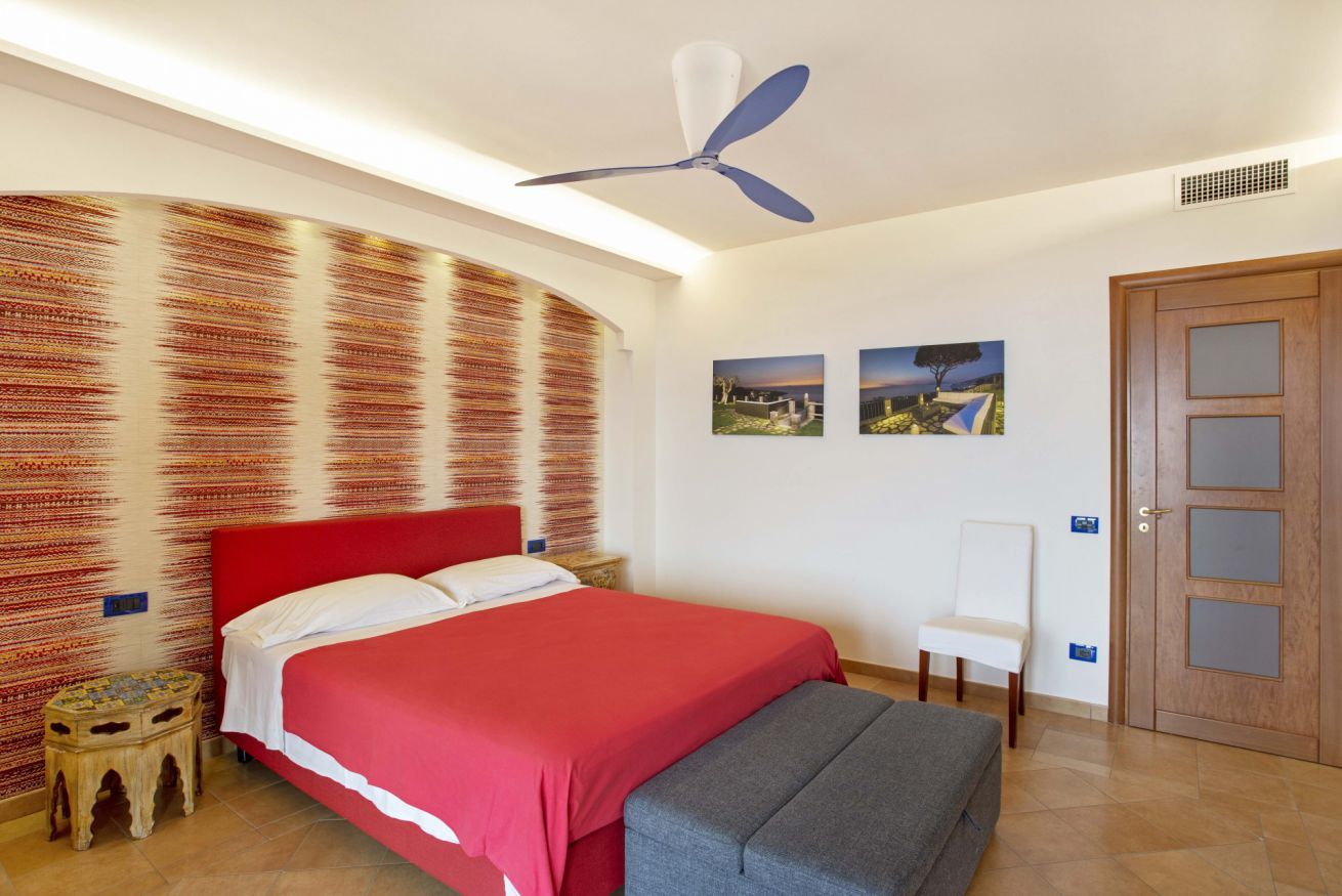 Un dormitorio con cama roja, ventilador de techo, puerta de madera y dos cuadros colgados en la pared.