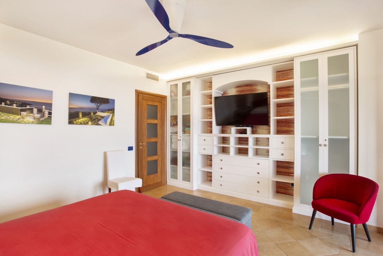 A bedroom with a bed, a red chair, a ceiling fan, a TV, and a wardrobe with glass doors.