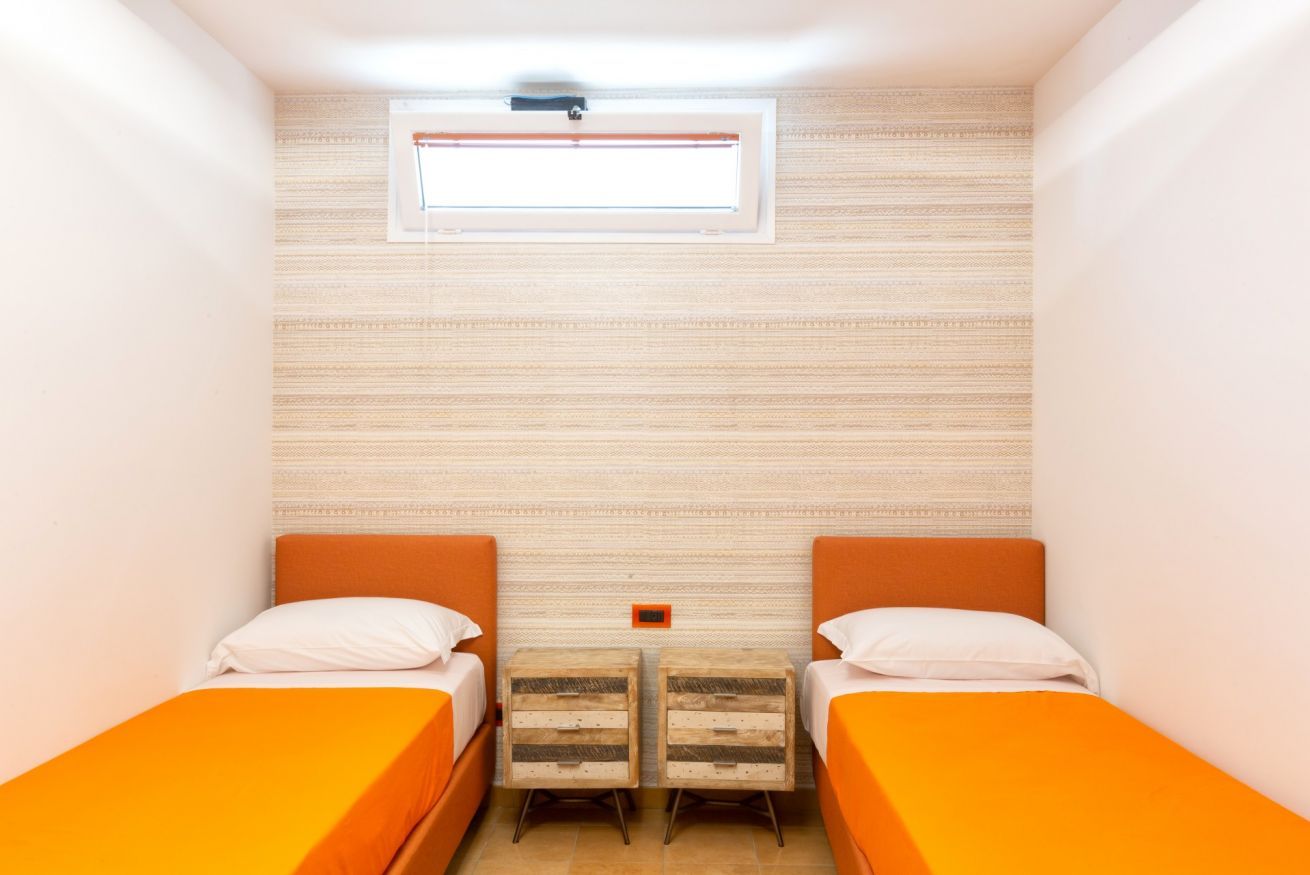 Una habitación pequeña e iluminada con dos camas, cada una con cabecero naranja y ropa de cama blanca. Hay una ventana sobre las camas, y una mesita de noche de madera con cajones entre ellas.