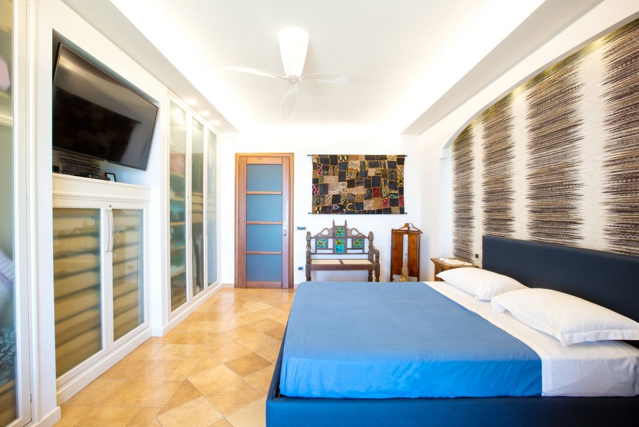 A bedroom with a bed, a ceiling fan, a TV, a wooden bench, and a wall with a textured pattern.