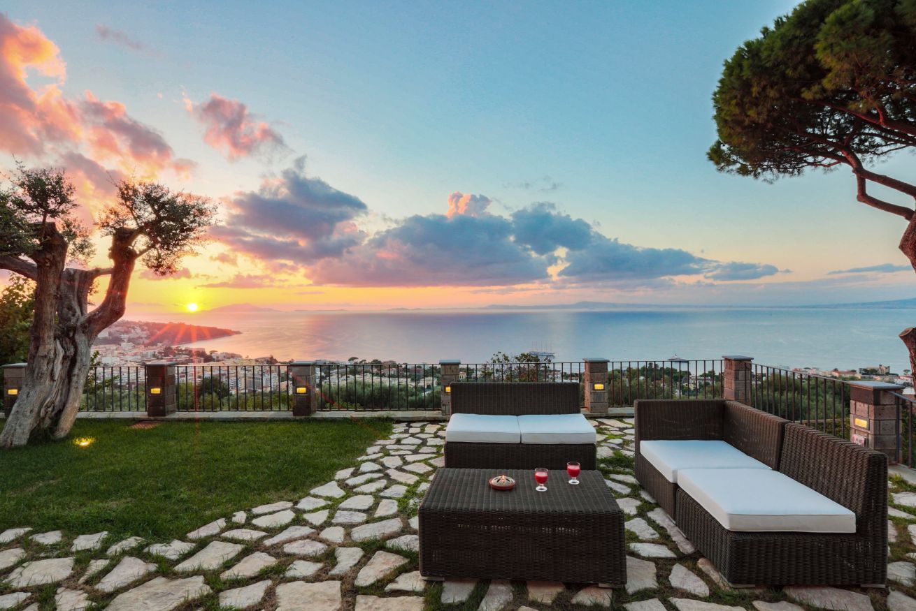An outdoor seating area with a view of the sunset over the sea, stone pathway, and a fence.