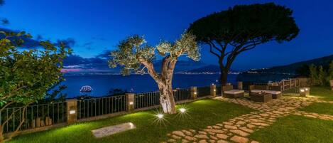 A nighttime outdoor area with a stone pathway, seating, and a view of the sea.