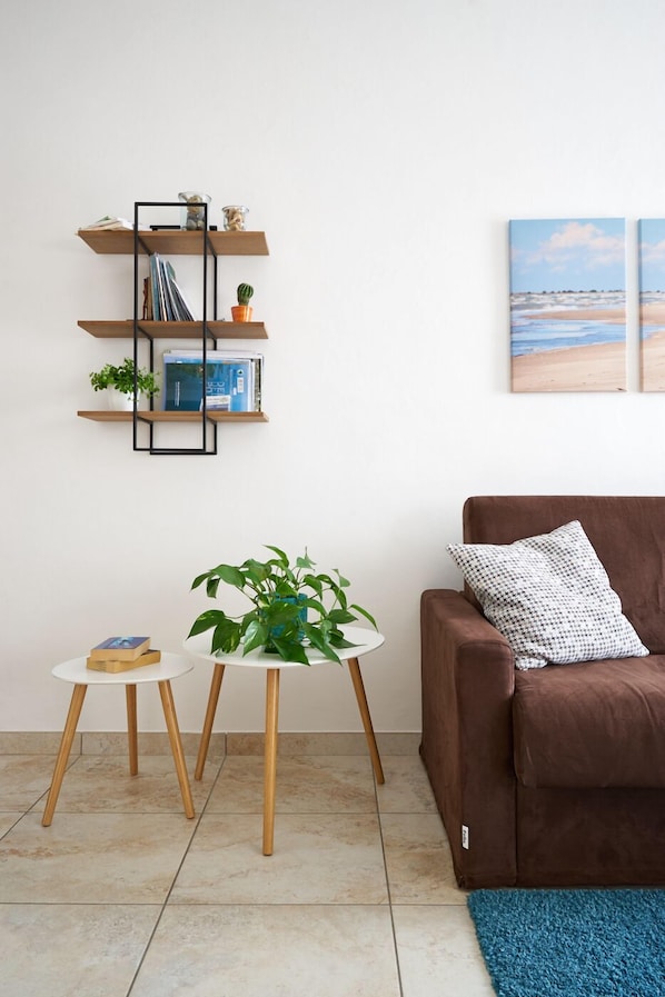 Un salon avec un canapé marron, une petite table blanche ornée d’une plante, une étagère en bois avec des livres et un cactus, et un tableau représentant une plage.