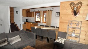 A cozy living room with a grey sofa, a wooden coffee table, and a dining area with a large wooden table and chairs. A kitchen is visible in the background with wooden cabinets and a window. - Typical chalet, combining the charm of wood and stone, overlooking the Isère river  (Tignes)