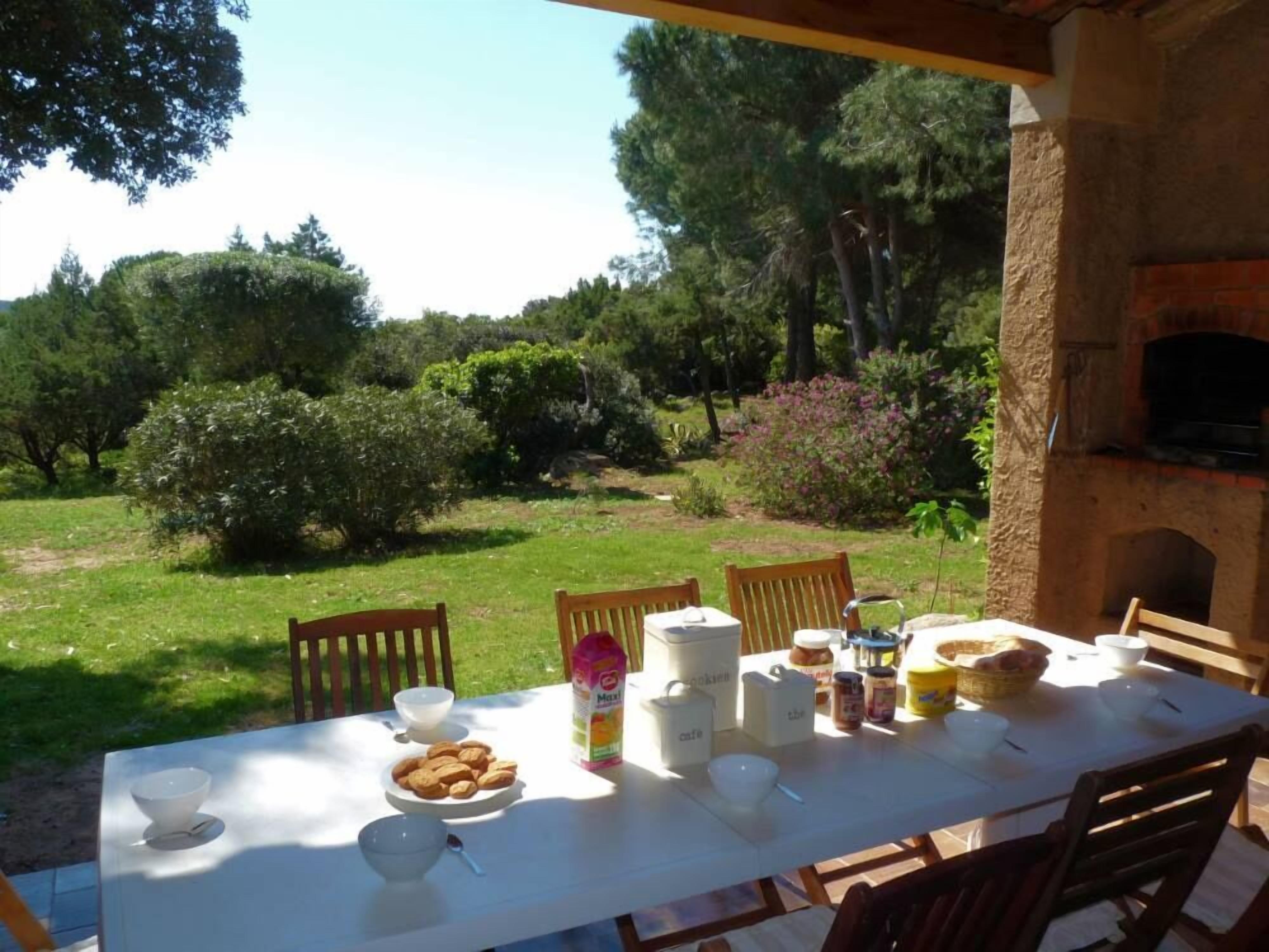 Una mesa de comedor al aire libre con diversos alimentos y condimentos, rodeada de sillas, y vista a un jardín y árboles en el fondo.
