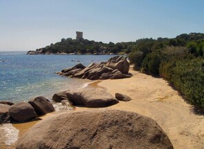 A beach with large rocks, a sandy path, and a tower on a hill in the distance.