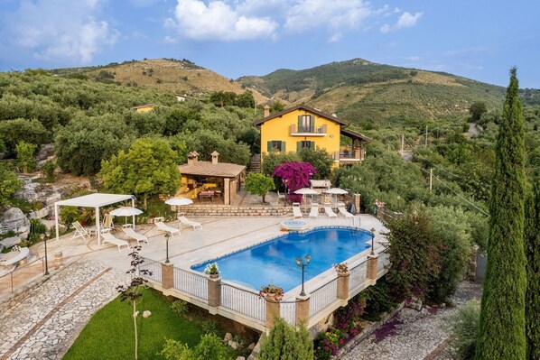 A yellow villa with a swimming pool, surrounded by lush greenery and hills.