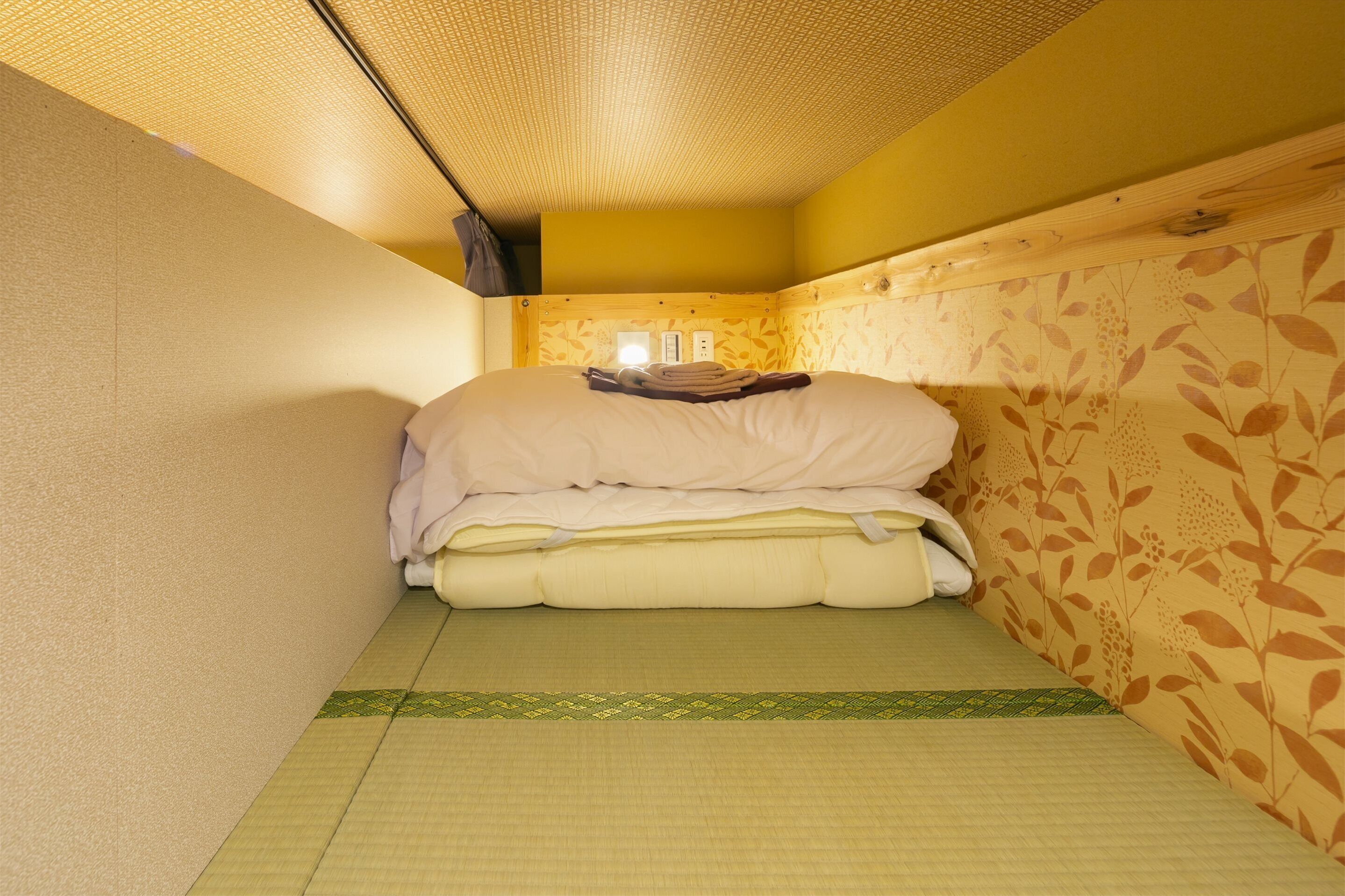 Une petite pièce étroite, avec un lit, une étagère fixée au mur et un papier peint à motifs.