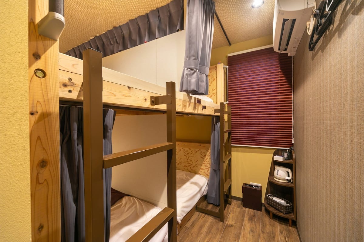 A bunk bed room with a wooden ladder, a small shelf with a kettle, and a window with blinds.