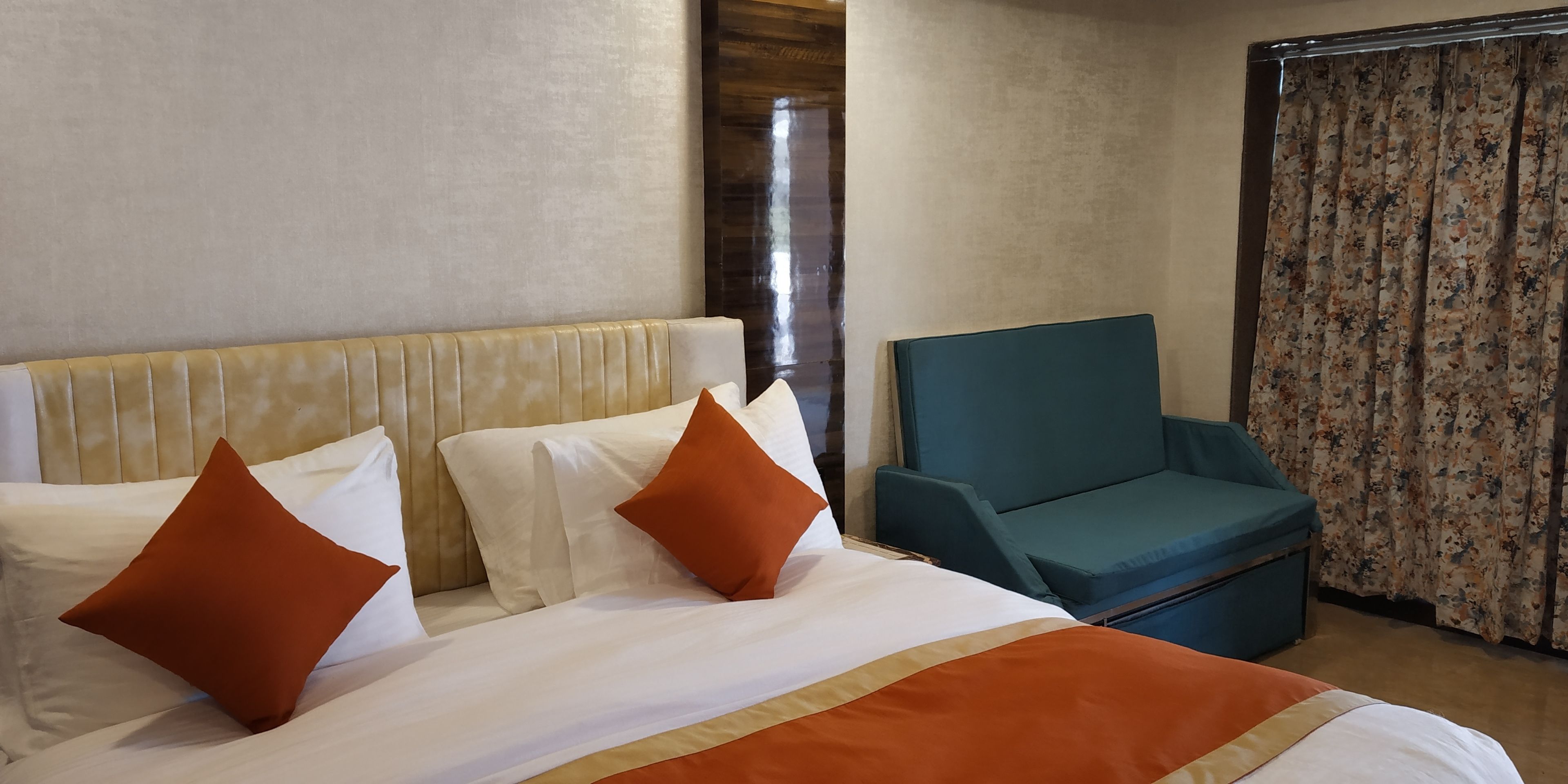 A hotel room with a bed, two pillows, a teal armchair, and a curtain.