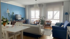 A modern living room with a white sofa, blue couch, and dining table. There are potted plants, a TV, and a lamp.