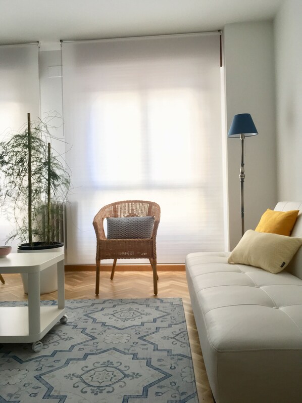 A living room with a white sofa, a wicker chair, a floor lamp, a patterned rug, and a potted plant.