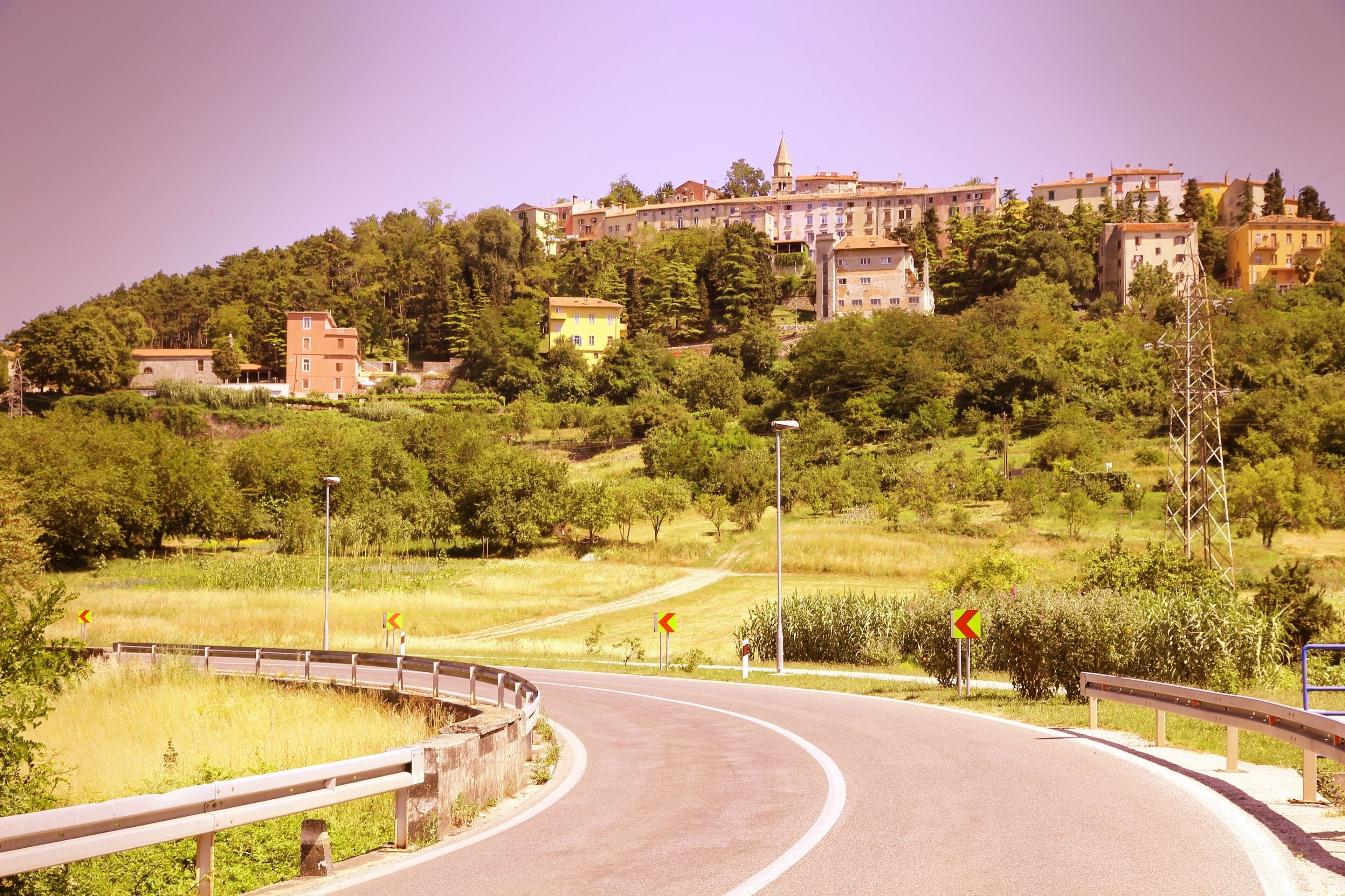 A winding road with a guardrail, leading to a hillside with buildings.