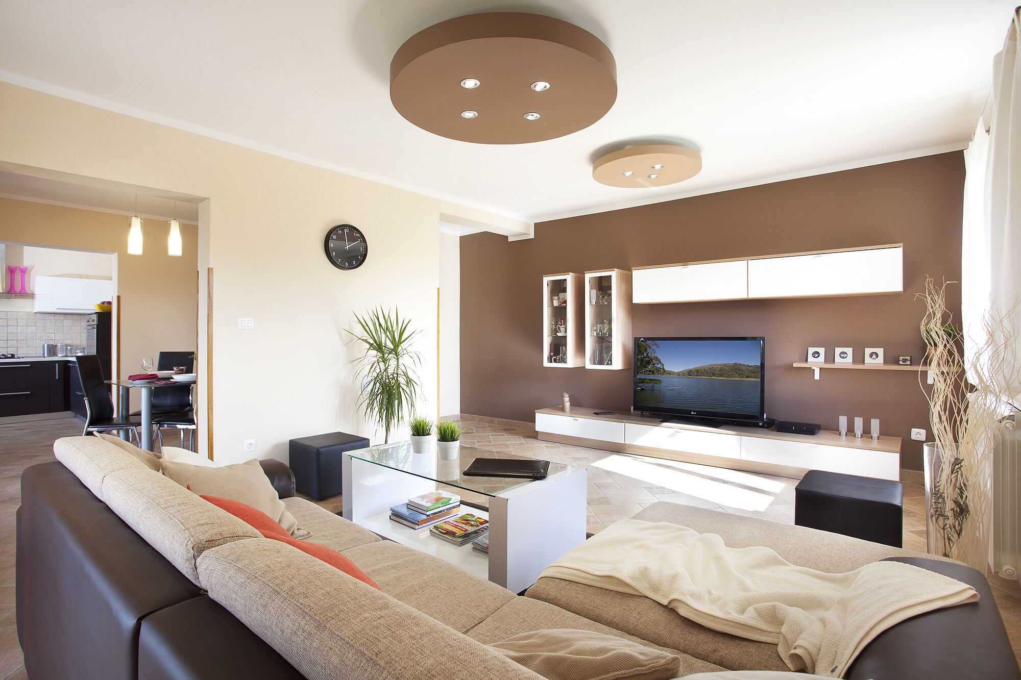 A modern living room with a large sofa, a coffee table, and a flat-screen TV. There is a kitchen area visible in the background.