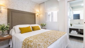 A bedroom with a bed, bedside lamps, a nightstand, a mirror, and a bathroom with a sink and towel rack. - Genteel Home Velarde II (Seville)