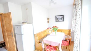Une petite pièce lumineuse avec une porte en bois, un réfrigérateur blanc, une table recouverte d’une nappe rose et deux chaises dotées de coussins à motifs floraux.
