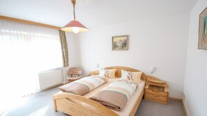 Une chambre à coucher avec un lit en bois, une table de chevet, une chaise, un radiateur et une fenêtre avec des rideaux.