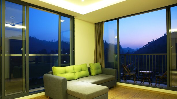 A modern living room with a grey sectional sofa, large floor-to-ceiling windows, and a balcony with outdoor furniture. - Casa Montana (Devikolam)