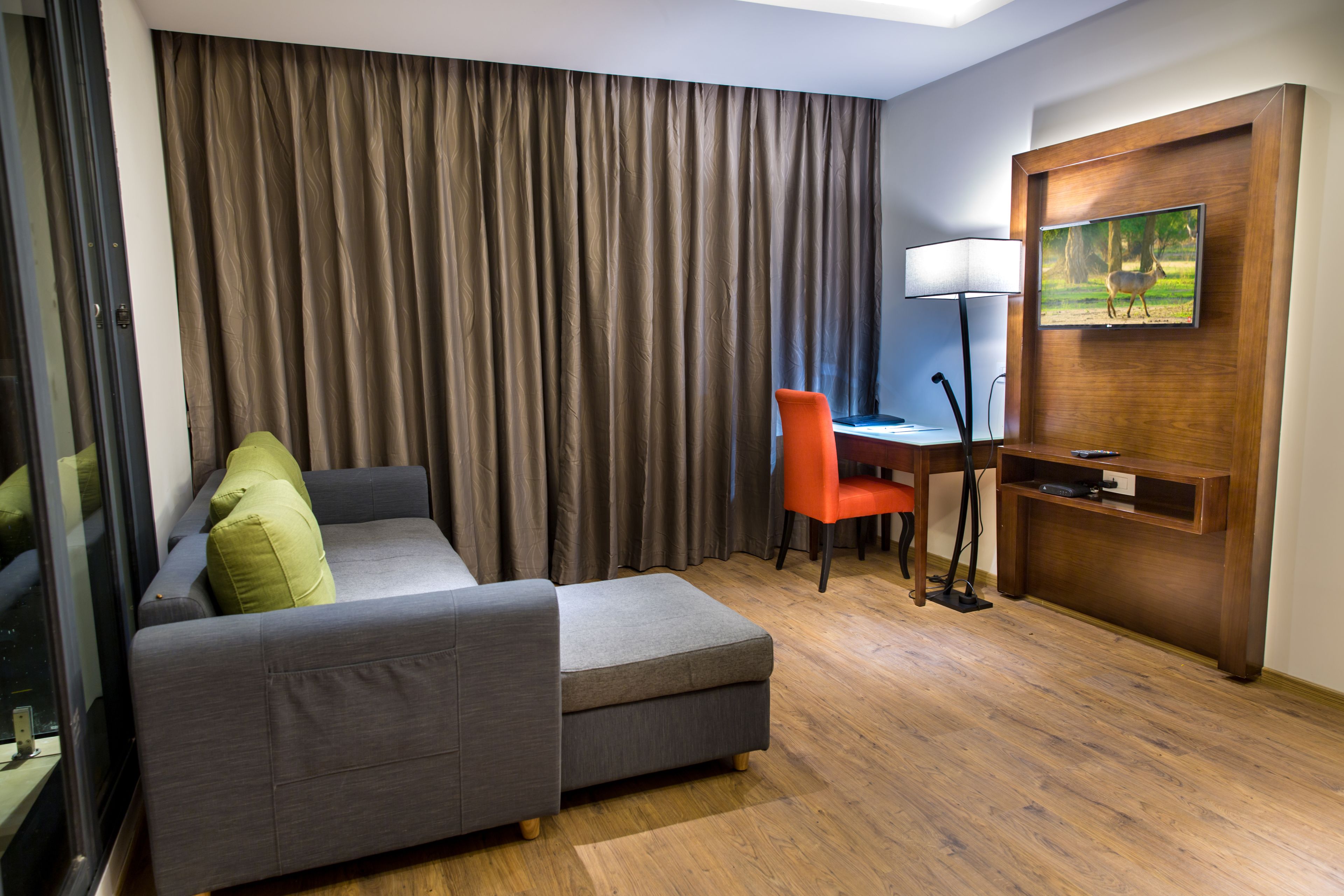 A modern hotel room with a grey sofa, an orange chair, a desk with a lamp, and a TV mounted on a wooden wall.