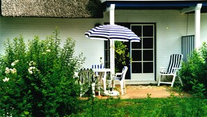A patio with a table and chairs, a striped umbrella, and a white chair. - Holiday apartment on the beach in a traditional thatched-roofed house  (Ostseebad zingst)