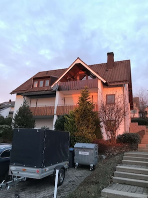A two-story house with a sloped roof, a balcony, and a chimney. There is a parked trailer and a dumpster labeled "Restmüll" in front of the house. - Designer apartment in the immediate vicinity of the bobsleigh run (Winterberg)
