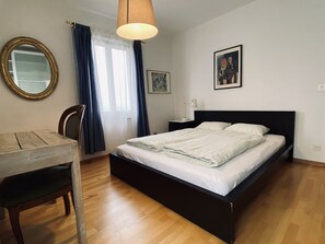 Une chambre à coucher avec un lit, un bureau, une chaise, un miroir et une fenêtre avec des rideaux.