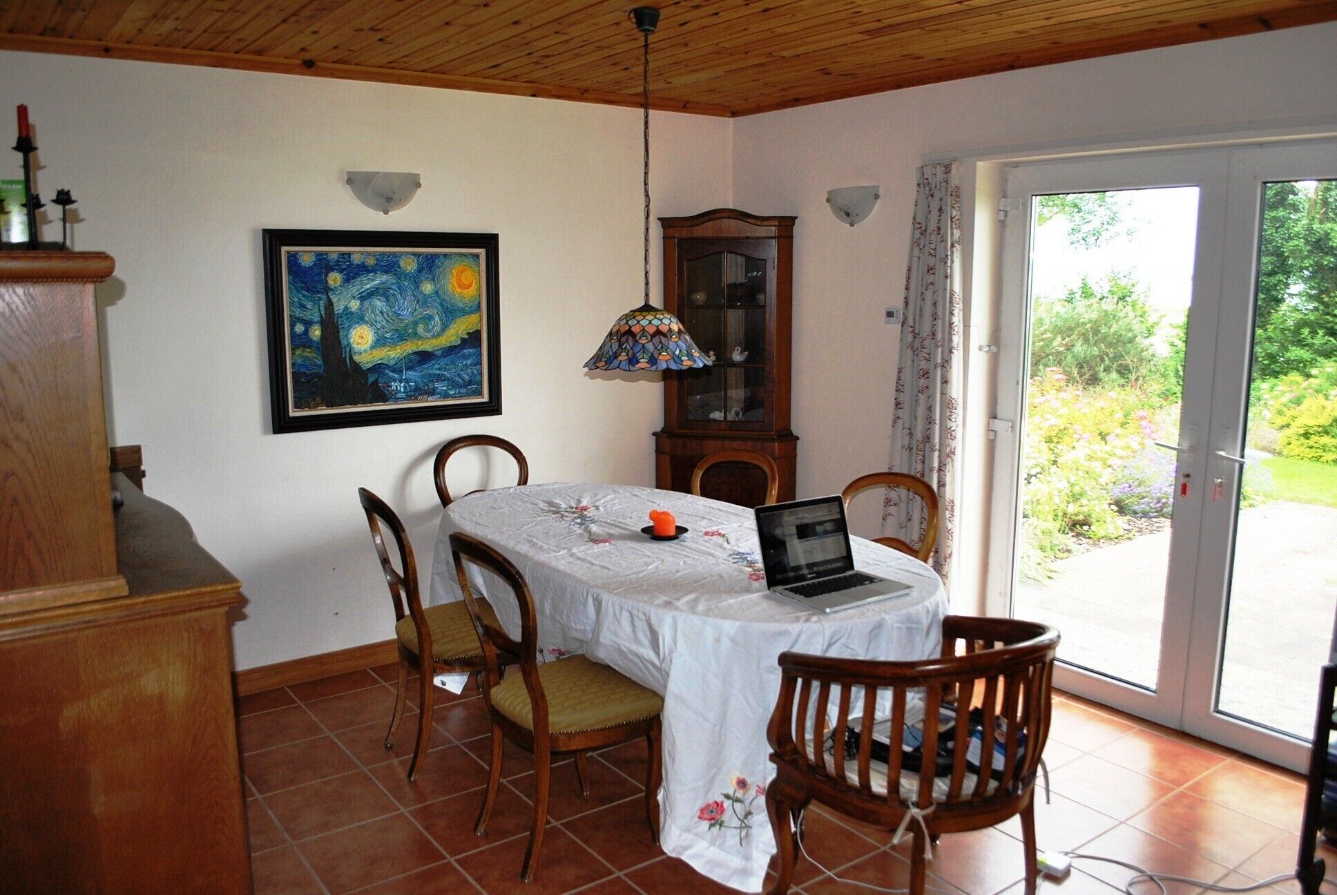 A dining area with a table, chairs, a laptop, a painting, and a cabinet.