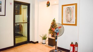 A hotel lobby with a glass door, a fan, a Christmas tree, a framed religious painting, and two fire extinguishers.