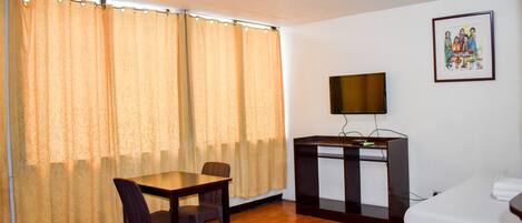 Una habitación con suelo de madera, una mesa con dos sillas, una cama, un televisor sobre un mueble bajo y una cortina.