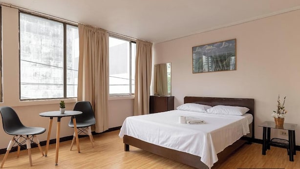 A hotel room with a bed, a small table with two chairs, a window with curtains, a nightstand, and a painting on the wall.