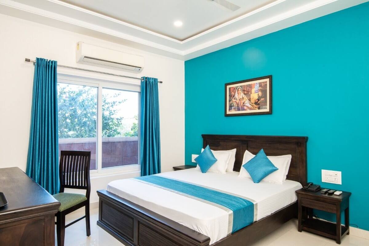 A bedroom with a large bed, a desk, and a chair. There is a window with curtains, an air conditioning unit, and a painting on the wall.