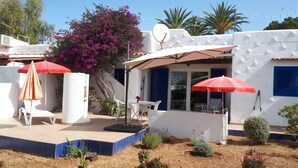 Un patio con paredes blancas, contraventanas azules y una puerta blanca. Hay dos sombrillas rojas, una silla blanca y una mesita.