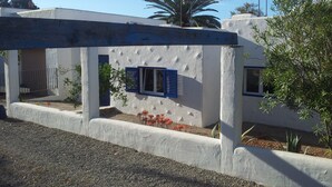 Un edificio moderno con fachada blanca y contraventanas azules, que cuenta con un balcón y una pequeña zona ajardinada con flores rojas.