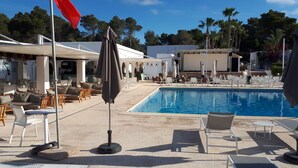 Zona junto a la piscina con sillones de descanso y sombrillas, una bandera y un edificio al fondo.